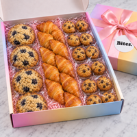 An open pastel gradient gift box filled with assorted baked goods, including croissants, blueberry muffins, and chocolate chip cookies, with the closed box and pink ribbon shown beside it on a marble surface.