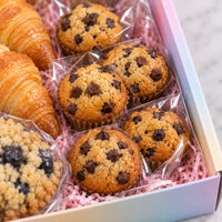 A gift box of assorted pastries on pink paper filler, including chocolate chip muffins, blueberry muffins, a croissant, and a crumb-topped pastry, each individually wrapped in clear cellophane.