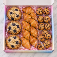 An open gift box filled with assorted baked goods, including blueberry muffins, blueberry oatmeal cookies, chocolate chip muffins, and croissants, arranged on pink shredded paper inside a pastel rainbow box.