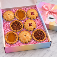 A gift box of seven mini fruit pies arranged in pink shredded paper, with an open view showing the golden crusts and two dark pecan pie centers. A pastel gradient gift box with a pink ribbon and the label 'Bites.