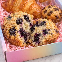 Blueberry muffins with crumb topping and a chocolate chip muffin in pink shredded paper inside a pastel gradient box, with a croissant in the background.