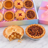 A boxed assortment of mini fruit pies displayed on a marble surface, with one apple pie cut open in front and a pink gift box in the background.