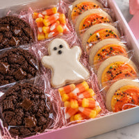 A boxed assortment of Halloween cookies and candy in a pink gift box. The assortment includes chocolate chip cookies, candy corn pieces, a ghost-shaped cookie, and sugar cookies topped with orange frosting and sprinkles.
