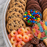 A close-up overhead view of a assorted snack tray filled with chocolate chip cookies, chocolate cookies, candy corn, M&M's candies, gummy bears, and gummy rings in a silver metal tray.