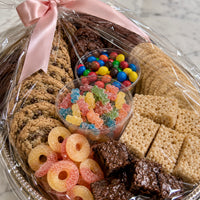 A wrapped snack assortment gift basket filled with cookies, crackers, candy, gummy bears, brownies, and a pink ribbon bow, photographed on a light marble surface.