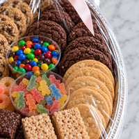 A large silver gift basket filled with assorted cookies, chocolate chips, candy corn, M&M's, gummy bears, and Rice Krispers, wrapped in clear plastic with a pink ribbon.