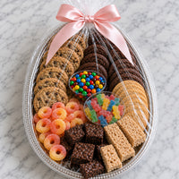 An Easter egg-shaped cookie and candy gift basket filled with assorted cookies, chocolate treats, gummy candies, and fruit loop candies, wrapped in clear cellophane with a pink satin bow, set on a white marble surface.