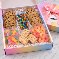 A gourmet cookie and candy gift box filled with chocolate chip cookies, colorful gummy candies, rice krisp treats, and sour gummy rings, displayed on a marble surface next to its pastel rainbow gift box.