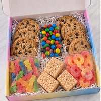 A boxed assorted cookie and candy gift set displayed from above in an open pastel gradient box. The set includes chocolate chip cookies, M&M's candies, gummy bears, square crackers, and pink ring-shaped gummy candies on white shredded paper.