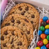 Three chocolate chip cookies arranged in white paper shred inside a gift box, next to a clear bag of colorful M&M's candies.