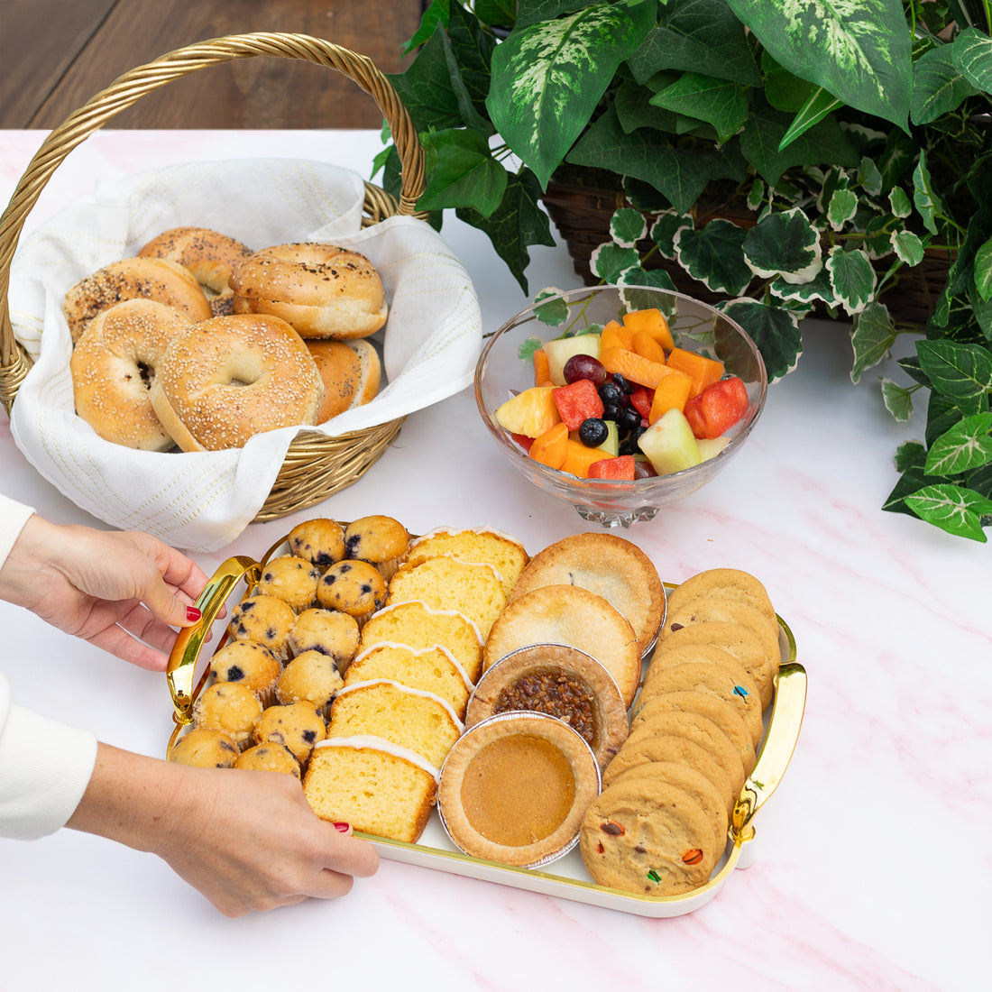 Assorted pastries on a tray with a basket of bagels and a bowl of fruit on a table.