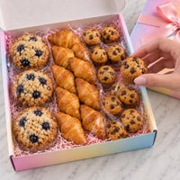 An open pastel gradient gift box filled with assorted baked goods, including blueberry muffins, chocolate chip muffins, and croissants, with a hand reaching for one muffin. A pink ribboned box sits beside it on a light marble surface.