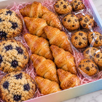 A gift box of assorted baked goods, featuring golden-brown croissants, blueberry muffins, chocolate chip muffins, and blueberry rice muffins arranged in pink shredded paper on a marble surface.