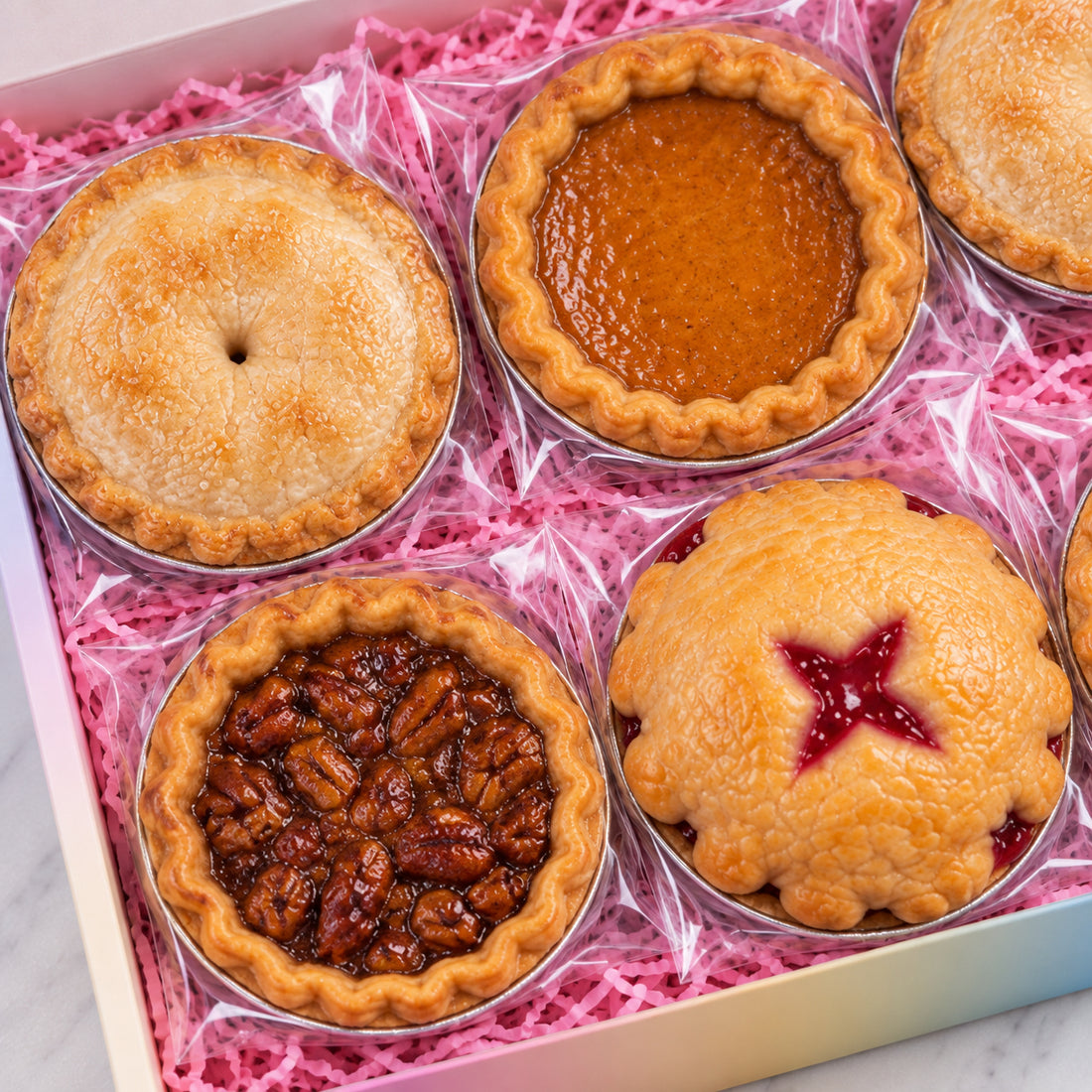 Six assorted mini pies and tarts arranged in a gift box lined with pink shredded paper. The pies include a lemon meringue pie, a pumpkin pie, a pecan pie, a berry pie with a star cutout, and a flaky pastry pie with a center pin.