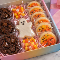 A boxed assortment of Halloween-themed cookies and candy in a pastel gradient box. The assortment includes chocolate chip cookies, a ghost-shaped cookie, orange and yellow decorated round cookies, and candy corn pieces, all individually wrapped in clear plastic over pink shredded paper.