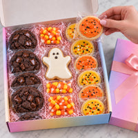 A Halloween-themed cookie gift box filled with assorted decorated cookies, including chocolate chip cookies, candy corn, a ghost cookie, and orange/yellow pumpkin cookies, shown with a hand holding one cookie. The box is open on a light marble surface, with a pink-lavender gift box and ribbon beside it.