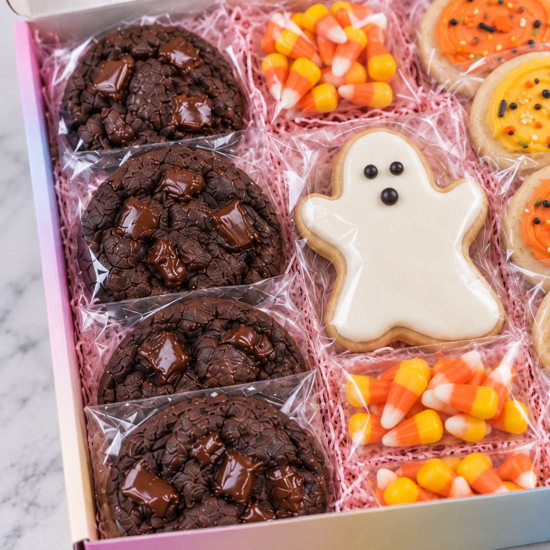A boxed assortment of Halloween cookies arranged in pink shredded paper. The set includes four chocolate chip cookies with chocolate chunks, candy corn bags, a ghost-shaped cookie with white icing and black eyes, and orange pumpkin cookies.