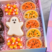 A boxed assortment of Halloween cookies displayed in a pastel box with pink shredded paper. The assortment includes a ghost-shaped cookie with white icing, orange frosted cookies with black and white sprinkles, candy corn cookies, and chocolate chip cookies.