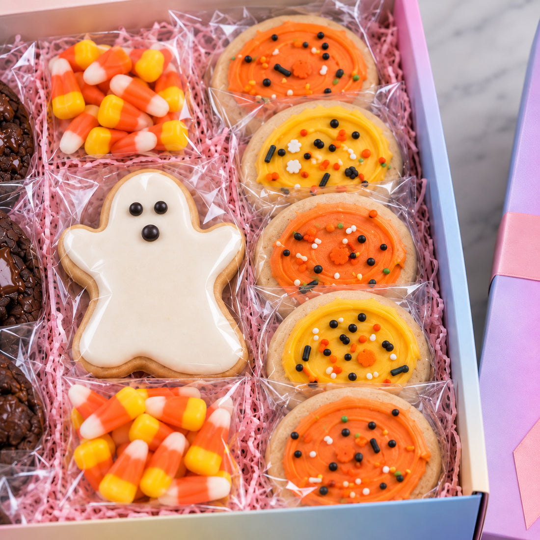 A boxed assortment of Halloween cookies displayed in a pastel box with pink shredded paper. The assortment includes a ghost-shaped cookie with white icing, orange frosted cookies with black and white sprinkles, candy corn cookies, and chocolate chip cookies.