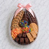 A gourmet cookie and candy gift basket arranged in an oval silver tray, wrapped in clear cellophane with a pink satin bow. The assortment includes chocolate chip cookies, chocolate brownies, Oreo cookies, cereal bars, candy corn, M&M's, and a bowl of colorful gummy candies, set on a white marble surface.