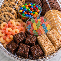 A large clear plastic basket filled with assorted baked goods and candy, including chocolate chip cookies, chocolate squares, Rice Krispers, M&M's, gummy bears, and orange rings, covered with clear plastic wrap.