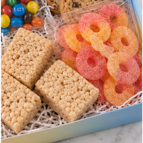 A boxed snack assortment featuring square rice crispy treats, bright pink and orange ring-shaped gummy candies, and a clear container of colorful M&M&