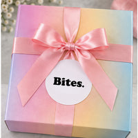 A pastel gradient gift box tied with a pink satin ribbon and bow, featuring a white circular label that reads 'Bites.' in black text.