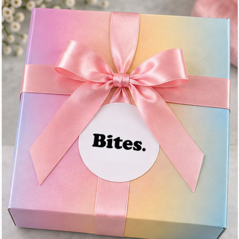 A pastel gradient gift box tied with a pink satin ribbon and bow, featuring a white circular label that reads &