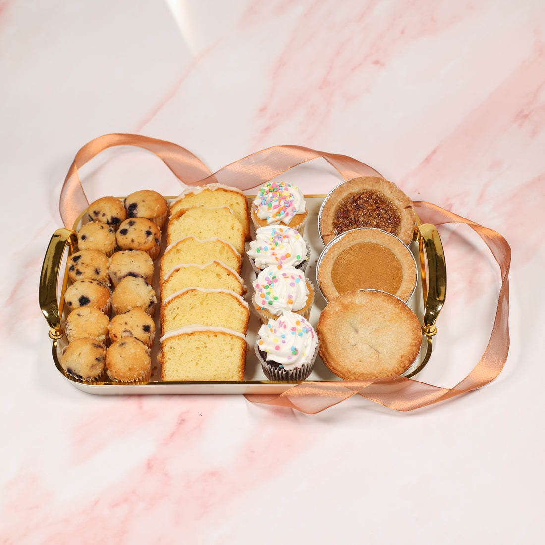 Assorted pastries including cookies, cakes, and cupcakes on a decorative tray with a pink background.